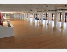 Großer, leerer Innenraum mit hellen Holzböden, weißen Säulen und einer hohen Decke mit Beleuchtungssystemen. Die Fensterreihen sind von roten Vorhängen eingerahmt, und der Raum wirkt offen mit minimaler Einrichtung.