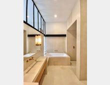 Modernes Badezimmer mit cremefarbenen Wänden und minimalistischem Design. Zu sehen sind eine eingelassene Badewanne, ein Waschbecken mit goldfarbenen Armaturen, ein großer Spiegel mit schwarzem Metallrahmen und integrierter Beleuchtung.