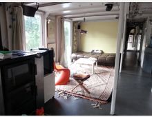 Wohnraum in einem hellen, offenen Bootshaus mit großen Fenstern, die auf eine grüne Landschaft blicken. Der Raum enthält einen Ofen, einen orangefarbenen Stuhl, einen kleinen Tisch, ein Bett im hinteren Bereich und einen Teppich auf einem dunklen Boden.