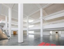 Großer, heller Innenraum eines modernen Gebäudes mit hohen weißen Säulen, mehreren offenen Ebenen und einer breiten Treppe mit dunkler Steinverkleidung. Der Boden besteht aus glänzendem Stein, und der Raum wird durch große Fensterflächen mit Tageslicht beleuchtet.