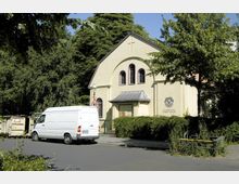Gelb gestrichene Kirche mit einfachem Giebeldach und drei schmalen, rundbogigen Fenstern an der Vorderseite, beschriftet als „Evangelische Ölberg-Kirche“. Im Vordergrund sind ein geparkter weißer Transporter, eine Müllmulde und umliegende Bäume sichtbar.