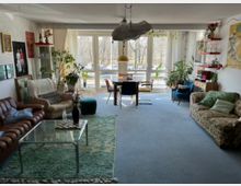 Bright, eclectic living room with large floor-to-ceiling windows and sheer curtains overlooking a street and trees. The space includes mixed vintage seating, a glass coffee table on a rug, a central dining table, wall art and shelves, and many potted plants throughout.