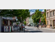 Straßenszene in einer kleinen Stadt mit mehrstöckigen, farbigen Häusern, Bäumen am Straßenrand und einem Gehweg mit Marktständen, die Kleidung verkaufen. Fußgänger und geparkte Autos sind zu sehen, während eine Bushaltestelle und ein Straßenschild den Bereich kennzeichnen.