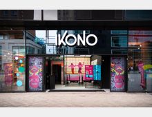 Die Abbildung zeigt die Eingangsfassade von "IKONO", einem modernen interaktiven Ausstellungsraum, mit großen Glasfronten und auffälliger dekorativer Gestaltung. Links und rechts des Eingangs befinden sich bunte, neonartige Kunstmotive, während im Innenraum eine beleuchtete Wand mit Figuren sichtbar ist.