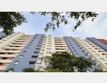 Hohe Wohnhausfassade mit mehreren Stockwerken, bemalt in Blau, Weiß und Rot, mit Balkonen an den meisten Wohnungen; im unteren Bereich sind Bäume und Äste zu sehen, die das Gebäude einrahmen. Der Himmel ist leicht bewölkt.