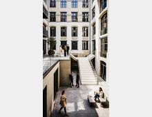 Innenhof eines modernen Bürogebäudes mit hellen Fassaden, großen Sprossenfenstern und dekorativen Ziegelbögen über den Fensterreihen. Eine breite, weiße Treppe und Glasgeländer verbinden zwei Ebenen; unten liegt ein gepflasterter Hofbereich mit Sitzbank und Pflanzkübel.