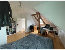 Dachzimmer mit Holzboden, schräger Decke und sichtbaren Holzbalken. Im Raum befinden sich ein Bett mit gemustertem Überwurf, ein Schreibtisch mit Computer, ein Regal voller Bücher und ein Sideboard unter einem kleinen Fenster.