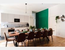 Moderne Wohnküche mit einem großen Esstisch aus Holz und acht braunen Stühlen, gedeckt mit Geschirr und Dekorationen. Im Hintergrund eine helle Einbauküche, eine grüne Wand und Dekorelemente wie Pflanzen an Wandregalen.