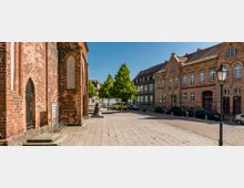 Außenansicht eines Platzes mit Pflastersteinen, links eine gotische Backsteinkirche mit Spitzbögen und Holztür, rechts ein Gebäude aus rotem Backstein beschriftet als „Evangelisches Gemeindezentrum“. Im Hintergrund stehen Bäume, parkende Autos und eine alte Glocke als dekoratives Element.