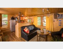 Gemütliches Wohnzimmer einer kleinen Holzhütte mit Holzverkleidung an Wänden und Decke. Schlichte Einrichtung mit schwarzen Ledersofas, gelben Vorhängen, einem runden Holztisch und einer kleinen offenen Küche im Hintergrund.