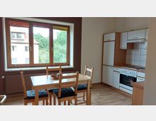 Helle Küche mit einem Essbereich, bestehend aus einem Holztisch und vier Stühlen. Rechts befinden sich Einbauküchenelemente mit Herd, Backofen und Fliesenspiegel; das große Fenster bietet Aussicht auf Bäume und umliegende Gebäude.