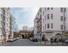 Straßenecke mit mehrstöckigen Wohnhäusern und geparkten Autos, im Hintergrund eine Ziegelbrücke mit einer S-Bahn darauf. Die Szene zeigt eine städtische Umgebung mit ruhiger Straßenatmosphäre und vereinzelten Bäumen.