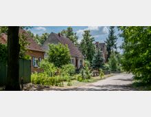 Eine ruhige Wohnstraße mit Einfamilienhäusern, die Ziegeldächer und gepflegte Vorgärten haben. Die Umgebung ist von Bäumen und Sträuchern gesäumt, während ein unbefestigter Weg entlang der Häuser verläuft.