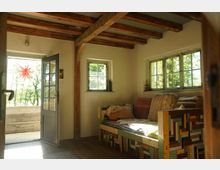 Gemütlicher Innenraum mit sichtbaren Holzbalken an der Decke, einem Sofa aus buntem Holz und Kissen, sowie zwei kleinen Fenstern, die viel Tageslicht einlassen. Eine geöffnete Tür führt zu einem Balkon mit Blick auf Bäume, dekoriert mit einem roten Sternanhänger.