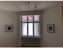 Ein heller Raum mit einer großen, weiß gerahmten Fensterfront, die nach außen auf ein Gebäude mit roten Dachziegeln blickt. An den weißen Wänden hängen zwei gerahmte Schwarz-Weiß-Fotografien, und unter dem Fenster befindet sich ein weißer Heizkörper.