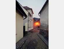 Schmale, gepflasterte Gasse in einer Wohngegend mit kleinen, eng beieinander stehenden Gebäuden mit hellen Fassaden und roten Ziegeldächern. Eine Straßenlaterne beleuchtet den Weg in der Dämmerung.