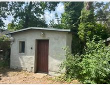 Kleines Garten- oder Geräteschuppen aus hellem Putz mit flachem Satteldach, roter Holztür und einem schmalen Fenster an der Vorderseite. Umgeben von dichtem Grün und hohen Bäumen, mit Buschwerk bis an die rechte Hauswand.