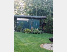 Garten mit gepflegtem Rasen, einer Feuerstelle im Vordergrund und einer blauen Holzhütte im Hintergrund. Blumen, Sträucher und ein kahler Baum schmücken den Bereich vor der Hütte, die von hohen Bäumen umgeben ist.