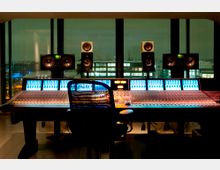 Tonstudio mit einem großen Mischpult, mehreren beleuchteten Kontrollanzeigen und mehreren Studio-Monitorlautsprechern, die vor einer Fensterfront platziert sind. Im Hintergrund ist eine Stadtlandschaft bei Nacht sichtbar.