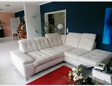 Wohnzimmer mit weißem Ecksofa auf weißem Marmorboden, rotem Teppich, Glas-Couchtisch mit Blumenarrangement und blauer Wand im Hintergrund. Links im Raum befindet sich ein Klavier vor einer rosa Wand sowie eine geöffnete Glastür mit Blick auf grüne Pflanzen.