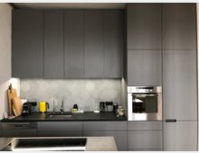 Moderne Küche mit matten, grauen Einbauschränken und einer Arbeitsplatte aus dunklem Material. Die Rückwand ist mit sechseckigen, grauen Fliesen gestaltet, und es sind ein Edelstahl-Spülbecken, ein Ofen, eine Spülmaschine und diverse kleine Elektrogeräte wie ein gelber Wasserkocher zu sehen.