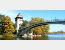 Ein historischer Wachturm mit der Aufschrift "Insel Berlin" steht am Ufer eines Flusses, umgeben von herbstlich gefärbten Bäumen. Eine gewölbte Fußgängerbrücke aus Metall führt darüber hinweg, während am Ufer kleine Boote festgemacht sind.