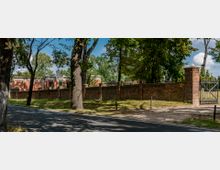 Straßenansicht einer historischen Backsteinmauer mit Toren, die ein Gelände mit Gebäuden im Hintergrund umgibt, umgeben von Bäumen und Vegetation. Die Szenerie zeigt eine ruhige Umgebung mit einer Asphaltstraße im Vordergrund.