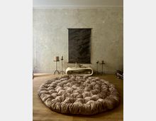 Ein minimalistischer Raum mit einer großen, runden, gepolsterten Matte in Beige auf einem Holzboden. Im Hintergrund eine strukturierte Wand, ein schwarzes Wandbanner mit Text, ein helles, geschwungenes Konsolentisch mit Deko-Elementen und zwei Kerzenständern mit Kerzen.