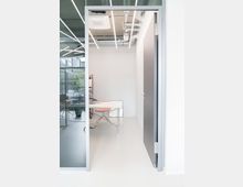 Ein moderner Büroraum mit glänzendem, hellem Boden, Glaswänden und sichtbarer Deckeninstallation mit Lüftungsrohren. Im Raum steht ein Schreibtisch mit einem rosa Stuhl, minimalistisch gestaltet.