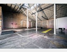 Weitläufiger, leerer Industrieraum mit freiliegenden Ziegelwänden, Metallträgern und einem Dach aus Wellblech. Im Vordergrund ist ein glänzender Betonboden zu sehen, rechts ein schwarzer Flügel, und links befindet sich eine erhöhte Bühne mit Stufen.