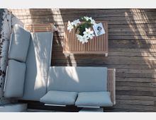 Terrasse mit Holzboden, darauf stehende Sitzmöbel mit hellgrauen Polstern und ein kleiner Holztisch, auf dem eine Vase mit weißen Blumen und ein Buch liegen. Links ist eine weiß gestrichene Wand mit Kletterpflanzen zu sehen.