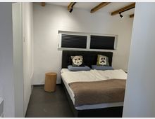 Modernes Schlafzimmer mit einem Doppelbett, schwarzem Kopfteil und dekorativen Kissen mit Gesichtermotiven. Die weißen Wände und die dunklen Bodenfliesen werden durch sichtbare Holzbalken an der Decke ergänzt; ein kleines Fenster mit dunklen Jalousien und ein runder Holzbeistelltisch runden das Design ab.