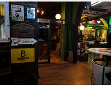 Innenraum einer irischen Pub-Bar mit dunkler Holzvertäfelung, vielen Vintage-Werbeschildern (u. a. Guinness und Bulmers) und warmen Kugellampen. Ein Durchgang führt zu einem Sitzbereich mit hohen Tischen, grün gestrichenen Bögen und beleuchteten Bar-Schildern an den Wänden.