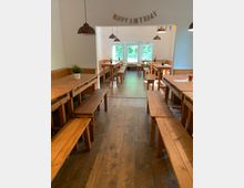 Ein heller Raum mit Holzbänken und -tischen, dekoriert mit einer "Happy Birthday"-Girlande und Wimpeln. Große Fenster im Hintergrund lassen Tageslicht herein, und Hängelampen aus Kupfer hängen von der Decke.