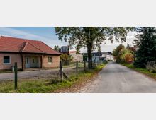 Straßenszene in einem ländlichen Dorf mit einem einzelnen Haus mit rotem Ziegeldach auf der linken Seite, einem Drahtzaun entlang der Straße und Bäumen, die teilweise die Straße säumen. Im Hintergrund sind weitere Gebäude und bunte Herbstbäume sichtbar.