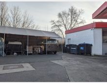 Eine Außenaufnahme eines Parkplatzes mit einer weißen Ziegelwand eines Gebäudes mit rotem Dachvorsprung rechts und einer Reihe von Müllcontainern daneben. Links steht ein überdachter Bereich mit einem alten Kleinbus und weiteren Gegenständen darunter, während kahle Bäume den Hintergrund säumen.