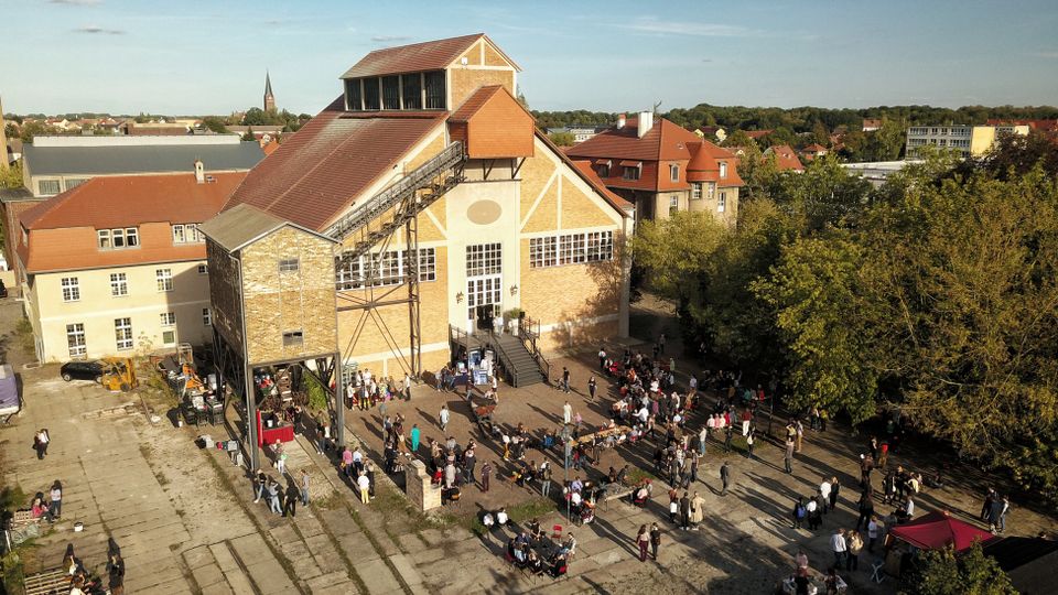 Luftaufnahme eines historischen Gebäudekomplexes mit Backsteinfassade und Satteldach, umgeben von Bäumen und niedrigen Häusern. Vor dem Hauptgebäude befinden sich zahlreiche Menschen bei einer Veranstaltung mit Tischen und Ständen im Freien.