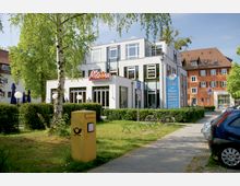 Ein zweigeschossiges, modernes Gebäude mit weißer Fassade, großen Glasfenstern und einem Schild „Meteora“ an der Vorderseite, das ein Restaurant und Café beherbergt. Im Vordergrund sind ein gelber Briefkasten, Büsche, Fahrräder und ein Gehweg zu sehen, umgeben von Bäumen; im Hintergrund steht ein traditionelles Gebäude mit roter Fassade.