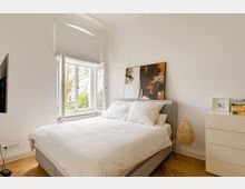 Helles Schlafzimmer mit einem großen Bett, einem offenen Fenster mit Blick auf einen Baum, einem abstrakten Gemälde an der Wand und einer weißen Kommode auf der rechten Seite. Der Raum hat Parkettboden und weiße Wände mit minimalistischer Einrichtung.