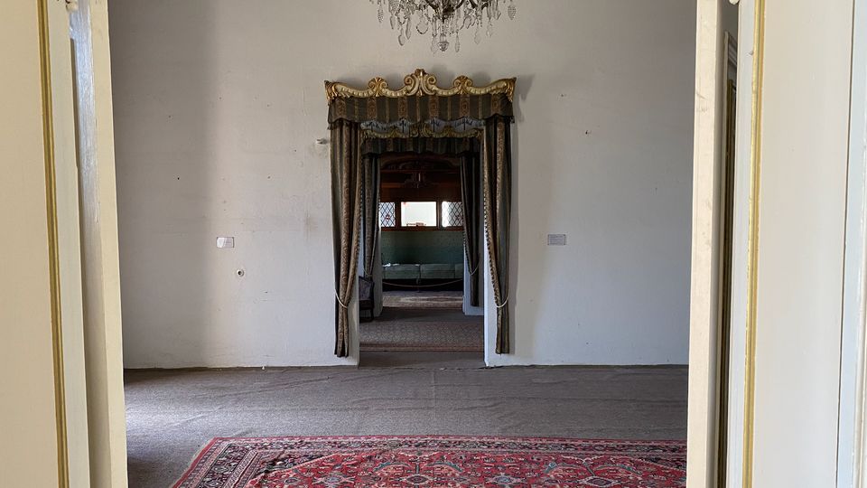 Blick in einen weitgehend leeren Raum eines historischen Gebäudes mit weiß verputzten Wänden, Kronleuchter und einem roten Orientteppich im Vordergrund. In der Mitte führt eine mit schweren Vorhängen und goldener Zierblende gerahmte Türöffnung in weitere Räume mit Fenstern.