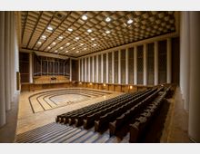 Innenansicht eines Konzertsaals mit hölzernem Boden und gestuften Sitzreihen. Im Hintergrund ist eine große Orgel eingebaut, die von Säulen und hochrechteckigen Wandelementen mit dekorativen Gittern eingerahmt wird, während die Decke aus einem Muster aus quadratischen Vertiefungen besteht.