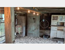 Innenansicht einer rustikalen Scheune mit groben Holzbalken, Kiesboden und einer hängenden Glaslaterne im Vordergrund. Im Hintergrund stehen alte Türen, ein Wagenrad an der Wand und ein weißer Schrank, darüber verläuft eine Lichterkette unter der Decke.