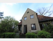 Ein Einfamilienhaus im traditionellen Stil mit einer Fassade aus beigefarbenem Putz, einem steilen Giebeldach mit roten Ziegeln und mehreren Fenstern, umgeben von Büschen und Bäumen in einem Garten. Der Eingangsbereich verfügt über eine dunkle Tür mit Glaseinsätzen.