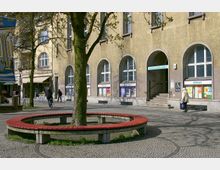 Öffentlicher Platz mit einem gepflasterten Boden, auf dem ein Baum steht, umgeben von einer runden roten Sitzbank. Im Hintergrund befindet sich ein Gebäude mit hellbrauner Fassade, großen Rundbogenfenstern und einem Eingang, der mit einem Schild „Bürgeramt“ gekennzeichnet ist.