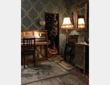 Ein gemütlicher Raum mit einem offenen Klavier, einer schwarzen Holzstuhllehne und einer Stehlampe mit auffälligem Stoffschirm. Im Hintergrund sind eine Wand mit dunkler Tapete und ein Türdurchgang zu sehen, der mit Lichterketten beleuchtet ist. Daneben befinden sich ein Spiegel, eine kleine Kommode, ein Verstärker und Musikinstrumente, darunter eine rote Gitarre.