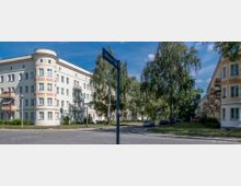 Mehrstöckige Wohngebäude im Stil früherer Jahrzehnte mit hellen Fassaden und abgerundeten Balkonen, die an einer baumbestandenen und gepflasterten Straßenecke liegen. Ein Straßenschild mit der Aufschrift "Saarlouiser Straße" steht im Vordergrund.