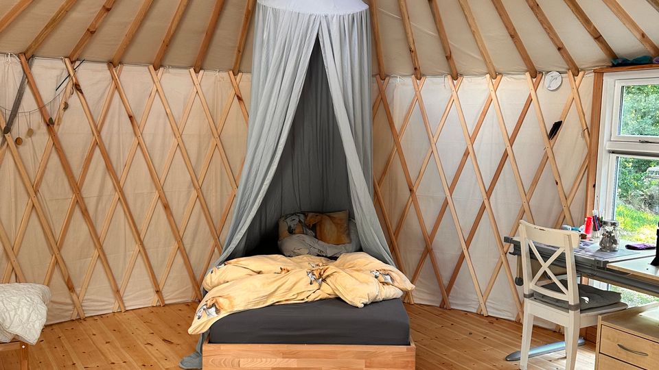 Innenansicht einer Jurte mit holzverkleidetem Boden und einem zentralen Oberlicht im Dach. In der Mitte des Raums steht ein Bett mit Baldachin, daneben ein Schreibtisch mit Stuhl und Fenster, das Tageslicht hereinlässt.