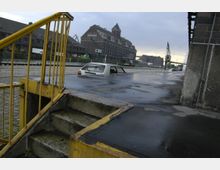 Industriehafen mit nassen Straßen und großen Backsteingebäuden im Hintergrund, ein teilweise überflutetes Auto auf der linken Seite und eine gelbe Metalltreppe im Vordergrund.
