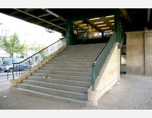 Treppe unter einer Hochbahnbrücke mit Metallgeländern führt zu einem Eingang mit Glasfenstern und beleuchtetem Innenraum. Im Hintergrund sind Straße, parkende Autos und Bäume zu sehen.