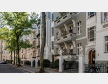 Straßenszene mit historischen Wohnhäusern im klassischen Stil, verziert mit Balkonen, Stuckornamenten und Skulpturen auf Torpfosten. Die Umgebung ist von Bäumen gesäumt, mit gepflasterten Gehwegen und parkenden Autos.